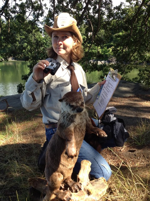 A woman wearing a hat, clutching a clipboard in one hand and binoculars in the other looks at the camera. A stuff river otter is in the foreground. 